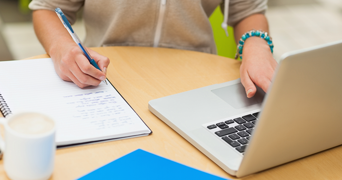 Student writing in a notebook and working on a laptop